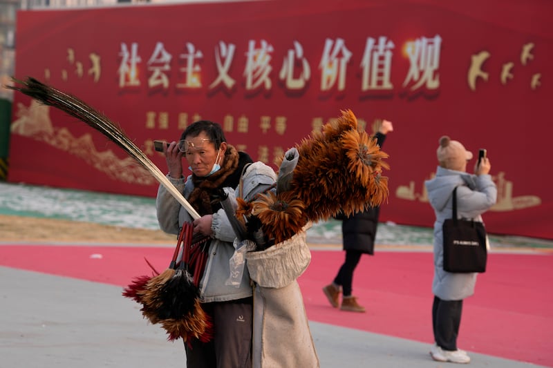 2024年1月20日,北京一名街头小贩在“社会主义核心价值观”宣传牌旁边一边听手机、一边兜售鸡毛掸子。
